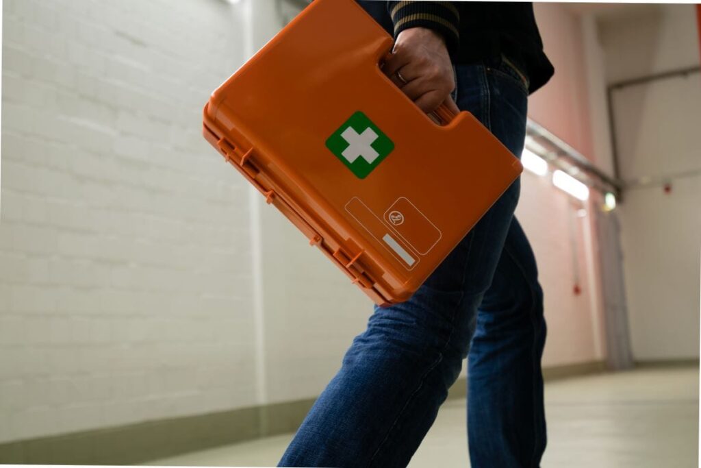 Ersthelfer im Betrieb – Beschäftigter trägt einen orangefarbenen Erste-Hilfe-Koffer